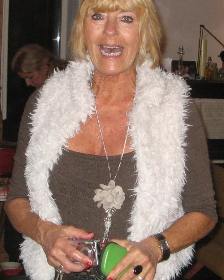 Femme blonde en chemise blanche, assise sur un canapé blanc, regardant par la fenêtre, pieds nus en sandales.