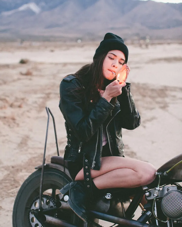 Femme en veste de cuir noire et bonnet, assise sur une moto dans un désert, allumant une cigarette.