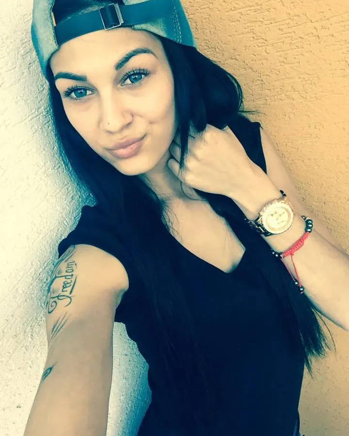 Femme latino portant une casquette renversée, un haut noir, un tatouage 'Freedom' sur le bras, regardant l'objectif.