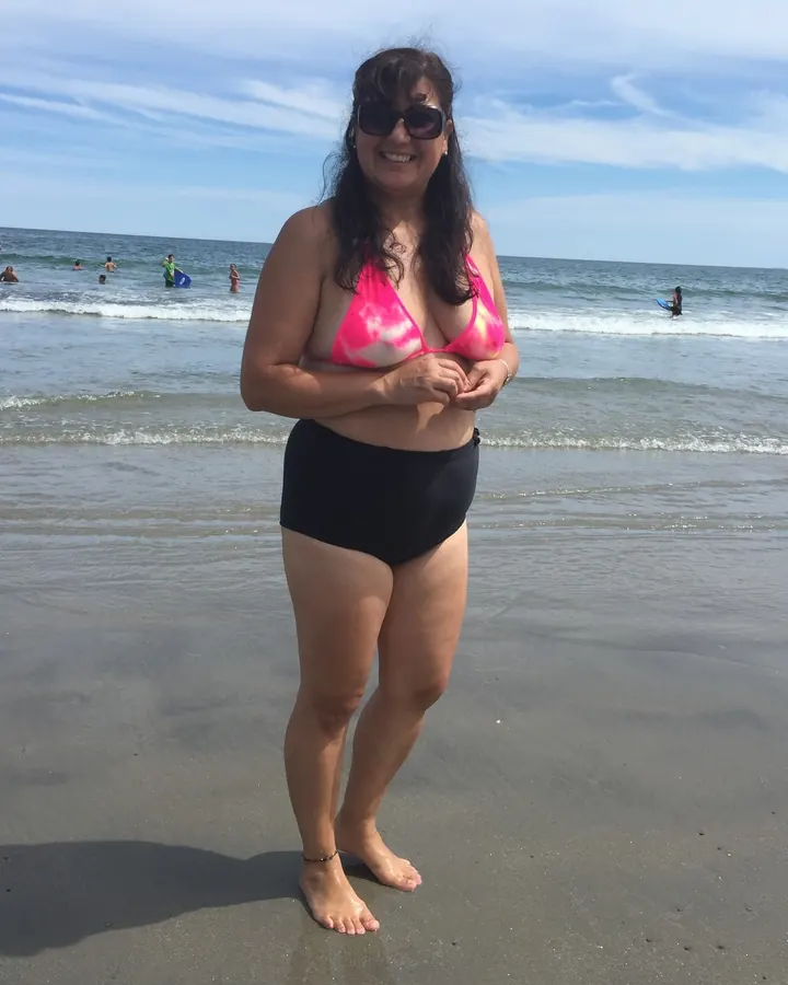 Femme souriante en maillot de bain rose et noir, debout sur une plage, devant la mer.