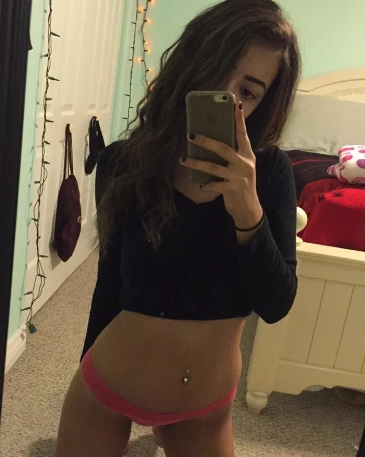 Femme brune en haut noir et sous-vêtements roses, agenouillée, prenant un selfie dans un miroir.