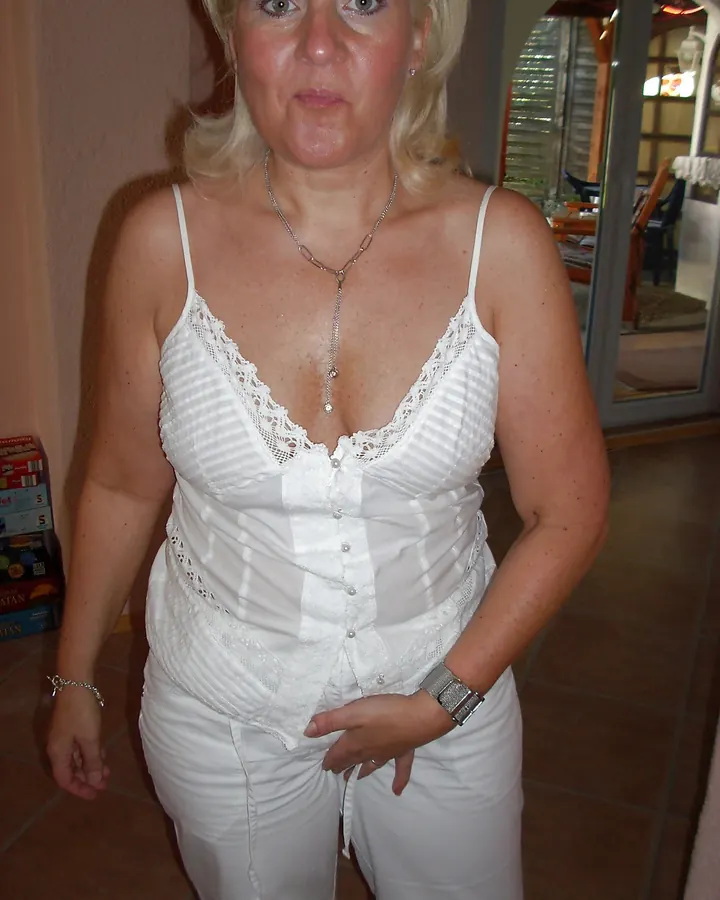 Femme blonde en chemisier blanc à lacets, pantalon blanc, collier long, regardant l'appareil, dans un intérieur avec carrelage au sol.