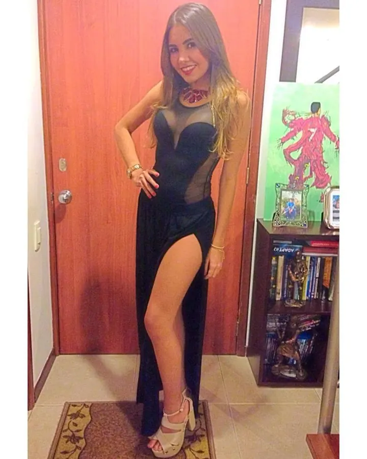 Femme aux cheveux châtains, en haut noir transparent, robe noire, talons noirs, debout dans une chambre, mains croisées devant elle.