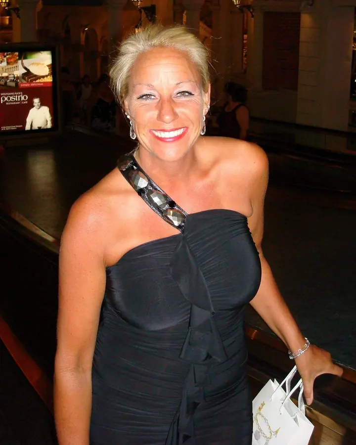 Femme blonde en robe noire asymétrique, souriante, tenant un sac blanc, dans un hall élégant avec un panneau publicitaire en arrière-plan.