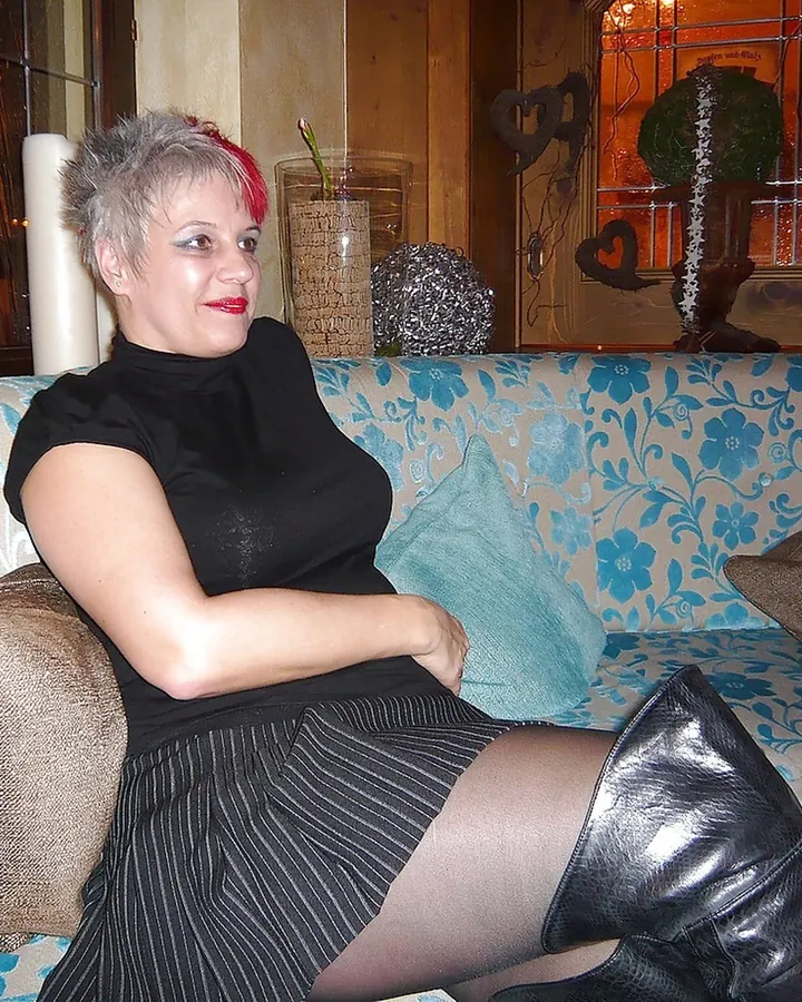 Femme aux cheveux gris courts, vêtue d’un haut noir et d’une jupe rayée, assise sur un canapé fleuri, les jambes croisées, bottes noires.