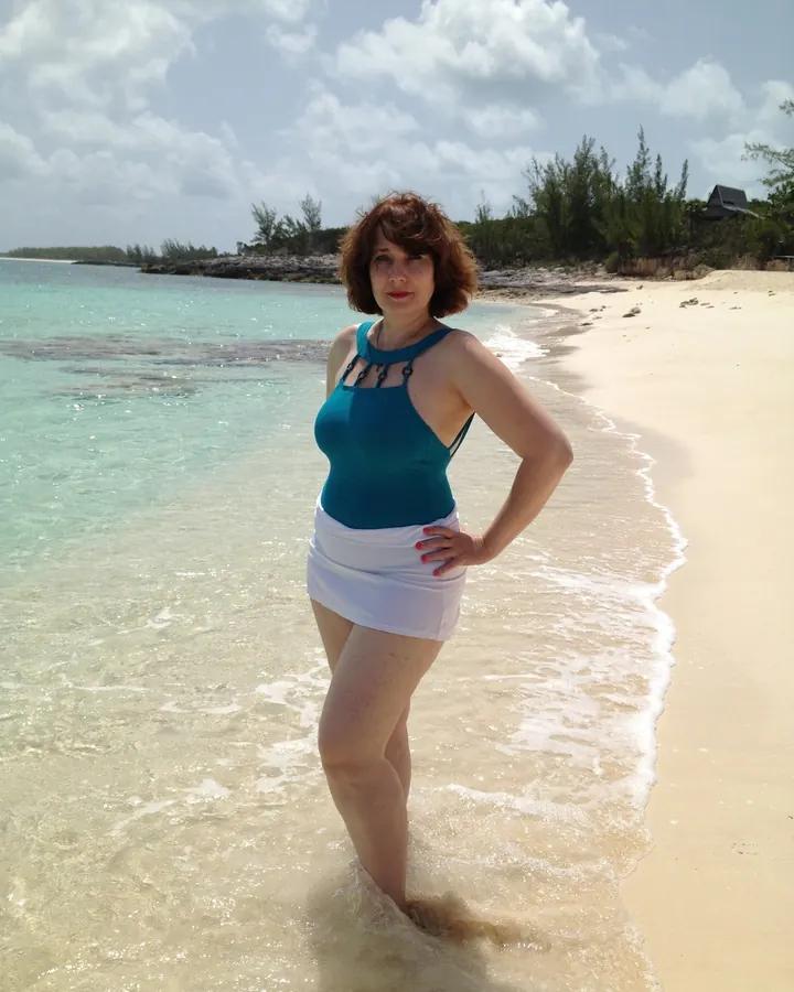 Femme aux cheveux châtains courts, en maillot de bain turquoise, debout dans l’eau claire d’une plage tropicale, main sur la hanche.