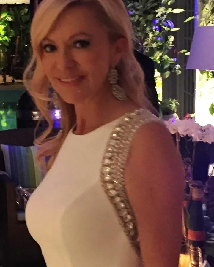Femme en robe blanche avec des strass dorés sur l'épaule, souriante, cheveux blonds, dans un décor nocturne.