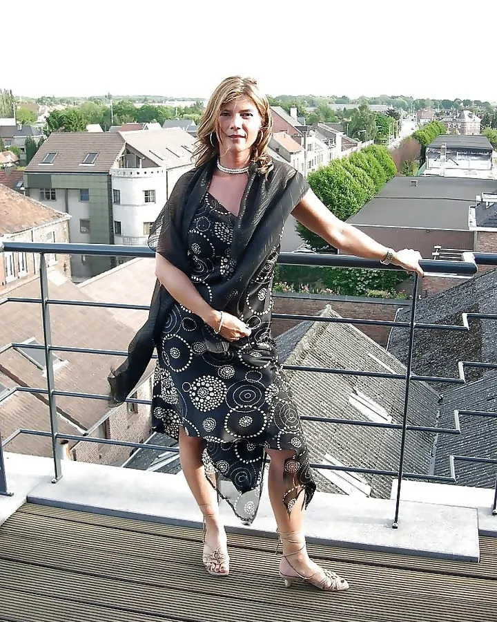Femme debout sur un balcon, en robe noire à motifs blancs, avec un foulard noir, regardant vers l'appareil.
