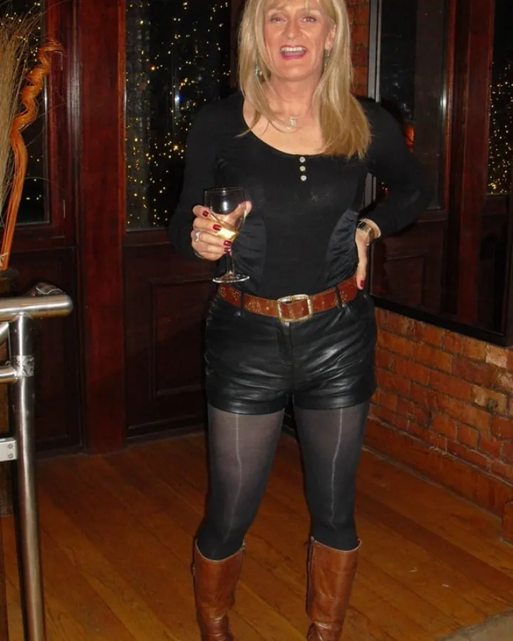 Femme blonde en robe noire en cuir, tenant un verre de vin, debout dans un hall décoré
