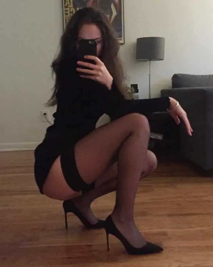 Femme aux cheveux bruns portant une robe noire courte, des collants noirs et des escarpins, agenouillée sur un parquet en bois dans un salon.