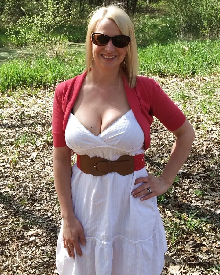 Femme blonde aux cheveux courts portant un haut rose de sport, assise en extérieur devant une maison avec des arbres en arrière-plan.