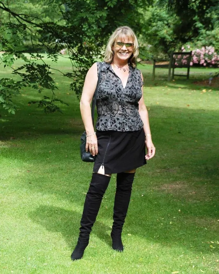 Femme blonde souriante, en tenue d’extérieur, debout dans un parc verdoyant, portant des lunettes de soleil et des bottes hautes.