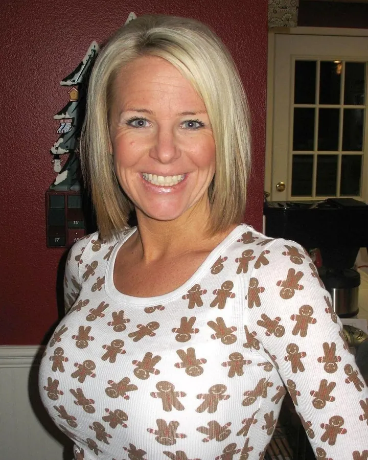 Femme blonde en haut blanc à imprimé de bonhommes, souriante, dans un intérieur chaleureux avec un décor rouge.