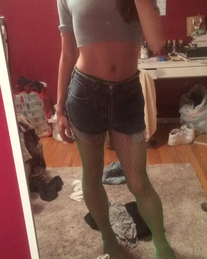 Jeune femme en haut gris et shorts en jean, debout devant un miroir dans une chambre, portant des collants verts à mailles.