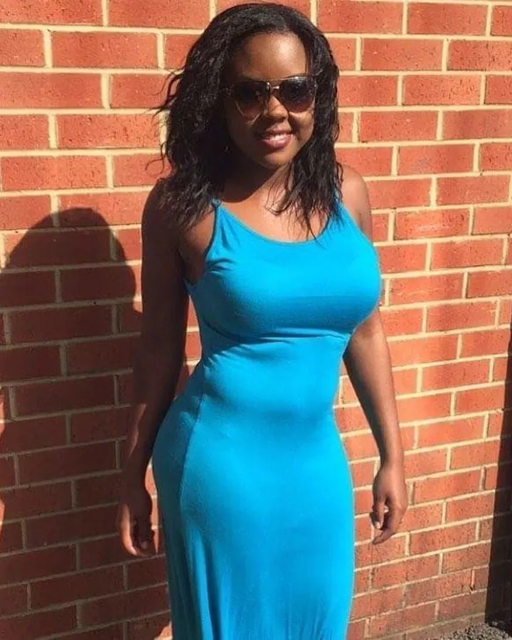 Femme noire en robe bleue ajustée, lunettes de soleil, souriant devant un mur de briques