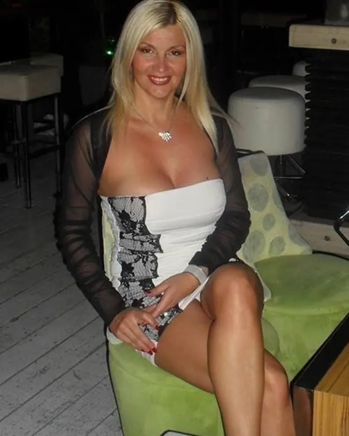 Femme blonde souriante, vêtue d'une robe blanche avec corset noir, assise sur un fauteuil vert