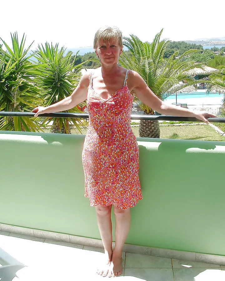 Femme européenne de 45 ans en extérieur, robe rouge ajustée, cheveux bruns mi-longs, regard direct, main sur hanche, appuyée contre porte en bois.