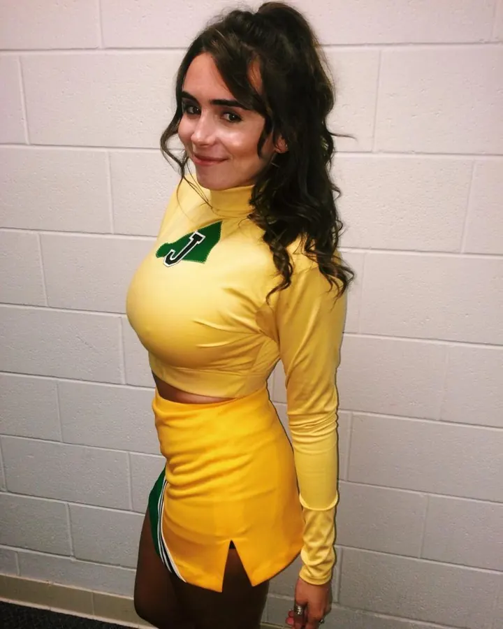 Femme européenne de 25 ans en extérieur, robe jaune moutarde ajustée, cheveux bruns longs, regard direct, pose de profil contre mur blanc.