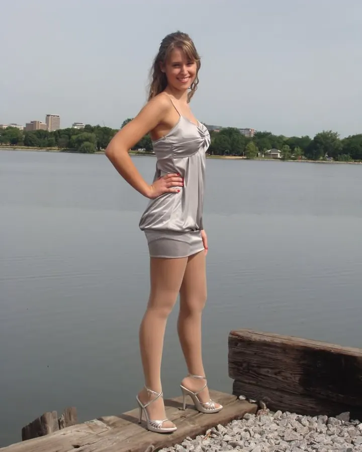 Femme européenne de 25 ans en extérieur, robe argentée courte, cheveux bruns ondulés, sandales argentées, sourire, pose sur jetée près d’un lac.