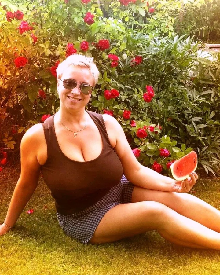 Femme aux cheveux gris courts, en haut marron et jupe à carreaux, assise sur l'herbe avec une tranche de pastèque, devant des roses rouges