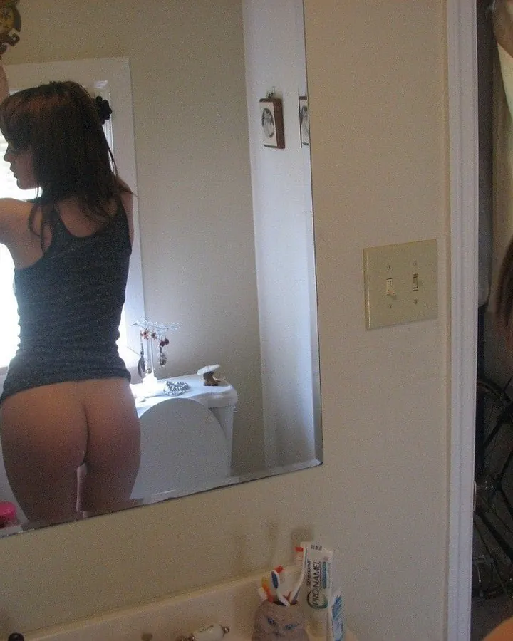 Femme aux cheveux bruns, en haut noir, prenant une photo de son reflet dans un miroir de salle de bain