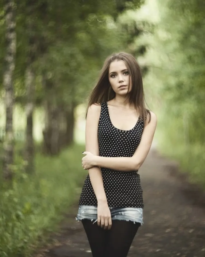 Jeune femme aux cheveux bruns debout dans une forêt, portant un haut noir à pois et un short en jean