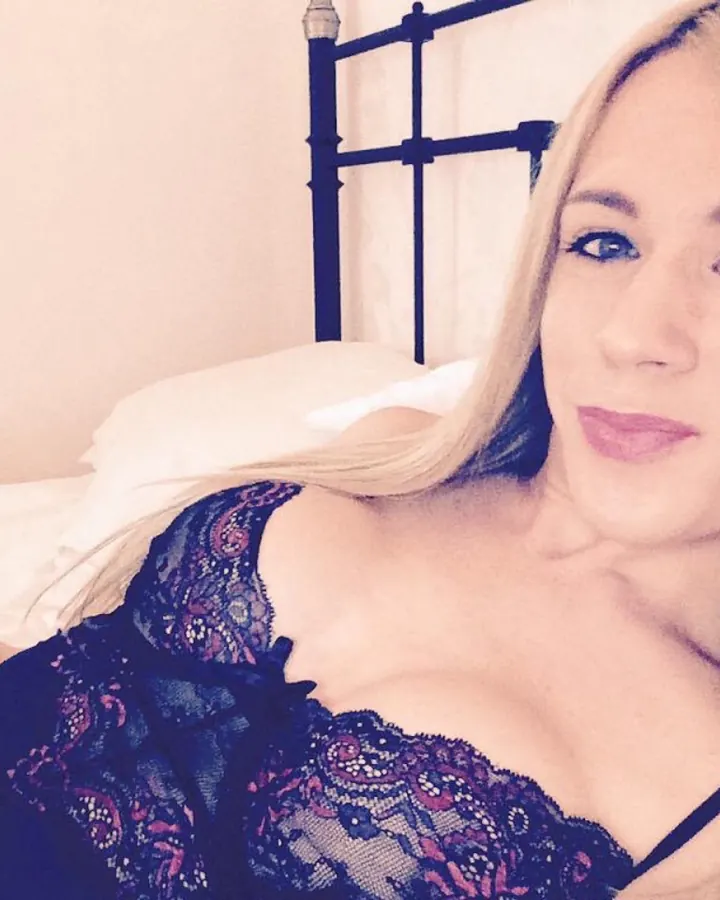 Jeune femme blonde en lingerie noire transparente, adossée à un mur clair, regardant l'appareil photo
