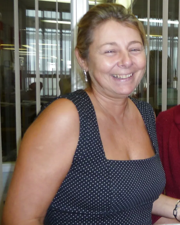 Femme aux cheveux blonds, en haut noir à manches longues, pantalon noir, assise sur des marches en béton, souriante
