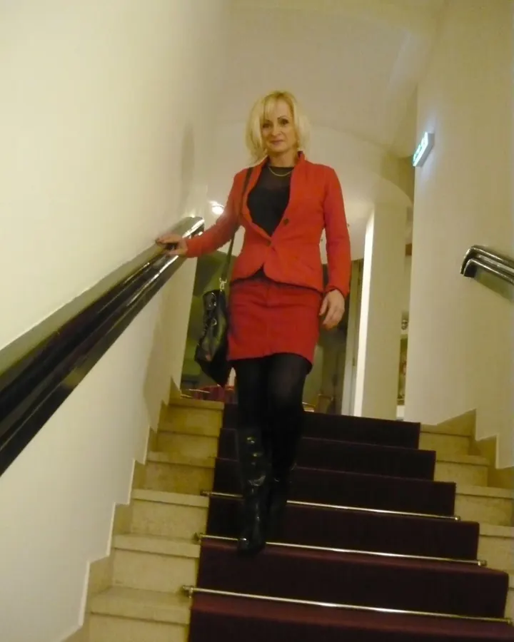 Femme blonde, en tailleur rouge, collants noirs, bottes hautes, descendant un escalier intérieur avec rampe métallique