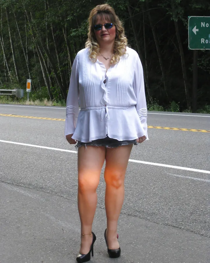 Femme aux cheveux blonds ondulés, en blouse blanche et shorts en jean, portant des lunettes de soleil, debout sur une route asphaltée