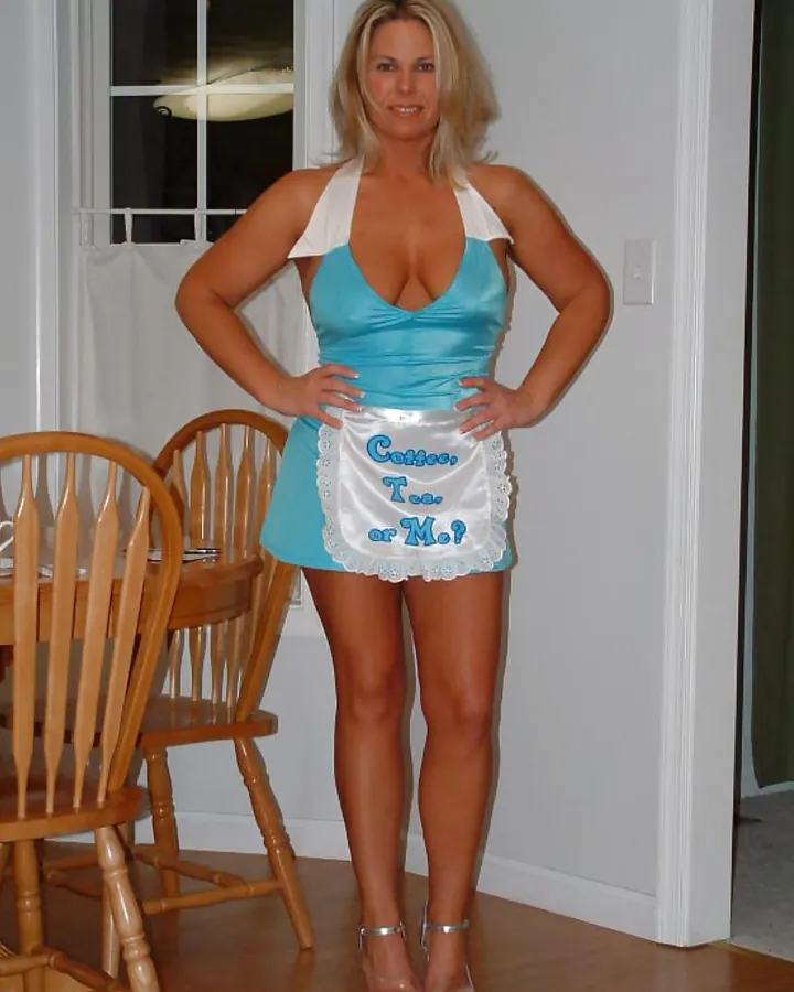 Femme blonde en robe bleue et tablier blanc avec inscription "Coffee, Tea or Me?", souriante, mains sur les hanches, dans une salle à manger.