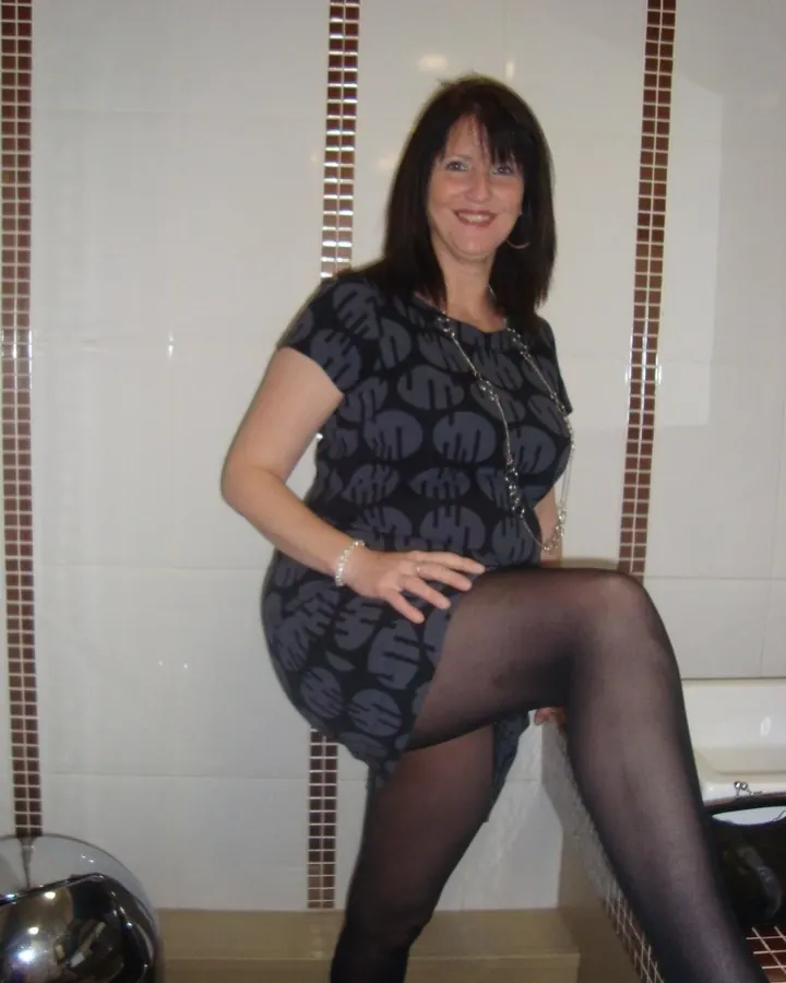 Femme en robe noire à motifs gris, assise sur un rebord de baignoire, les jambes croisées, les cheveux bruns mi-longs, les collants noirs.