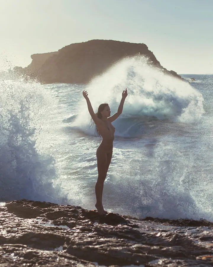 Femme nue, debout sur des rochers, les bras levés, face à une vague qui se brise derrière elle, sous un ciel clair.