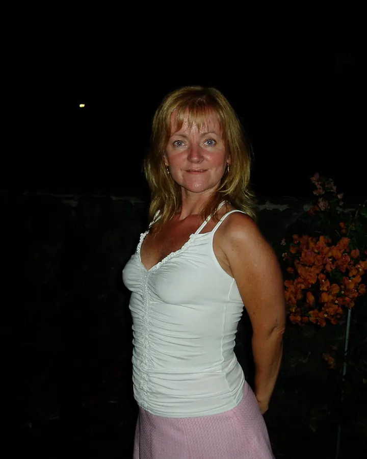 Femme en haut blanc et jupe rose, debout de nuit devant des fleurs orange, les cheveux blonds mi-longs.