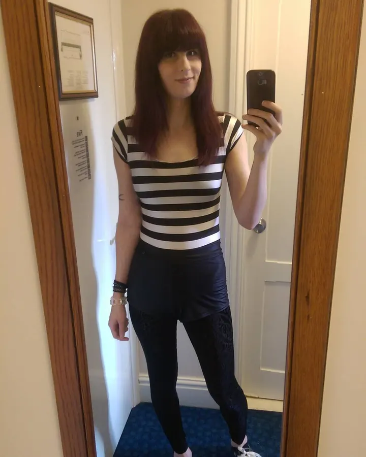 Femme aux cheveux longs roux, en haut à rayures horizontales noires et blanches, pantalon noir, posant devant un miroir, tenant un téléphone, regardant l'appareil.