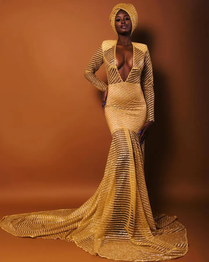 Femme africaine aux cheveux noirs courts, en robe dorée longue à sequins, avec capuchon assorti, posant contre un fond orange, main sur la hanche, regardant l'appareil.