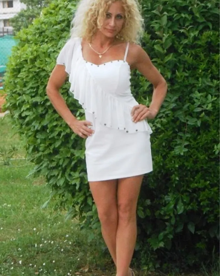 Femme blonde aux cheveux bouclés, en robe blanche courte avec volants, posant devant un buisson vert, chaussures à talons, mains sur les hanches, regardant l'appareil.
