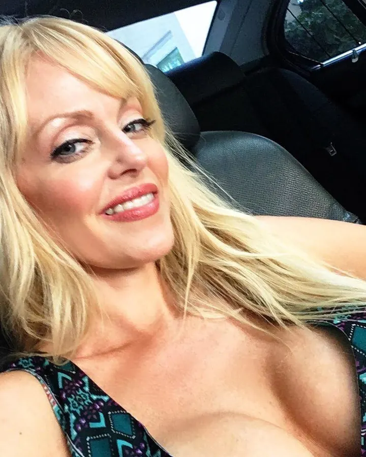 Femme blonde aux cheveux mi-longs, souriante, assise dans une voiture, portant un haut sans manches à motifs géométriques bleus et violets, regard tourné vers la caméra.
