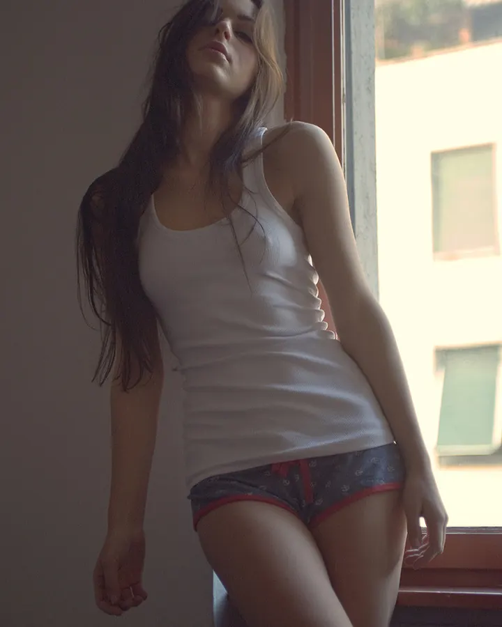 Femme aux cheveux bruns portant un soutien-gorge noir et un string, debout près d'une fenêtre avec vue sur une ville.