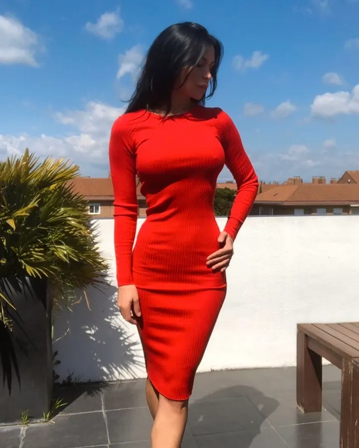 Femme aux cheveux bruns-noirs, en robe rouge ajustée, posant sur un balcon ensoleillé.
