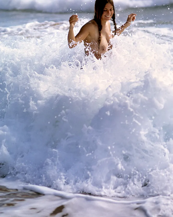 Femme aux cheveux châtains, souriante, les seins nus, émergeant d'une vague à la plage.