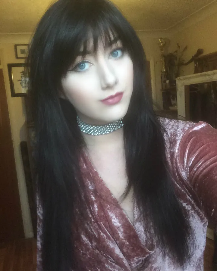 Femme aux cheveux noirs, regard bleu, en robe velours bordeaux, collier strass, prenant un selfie.