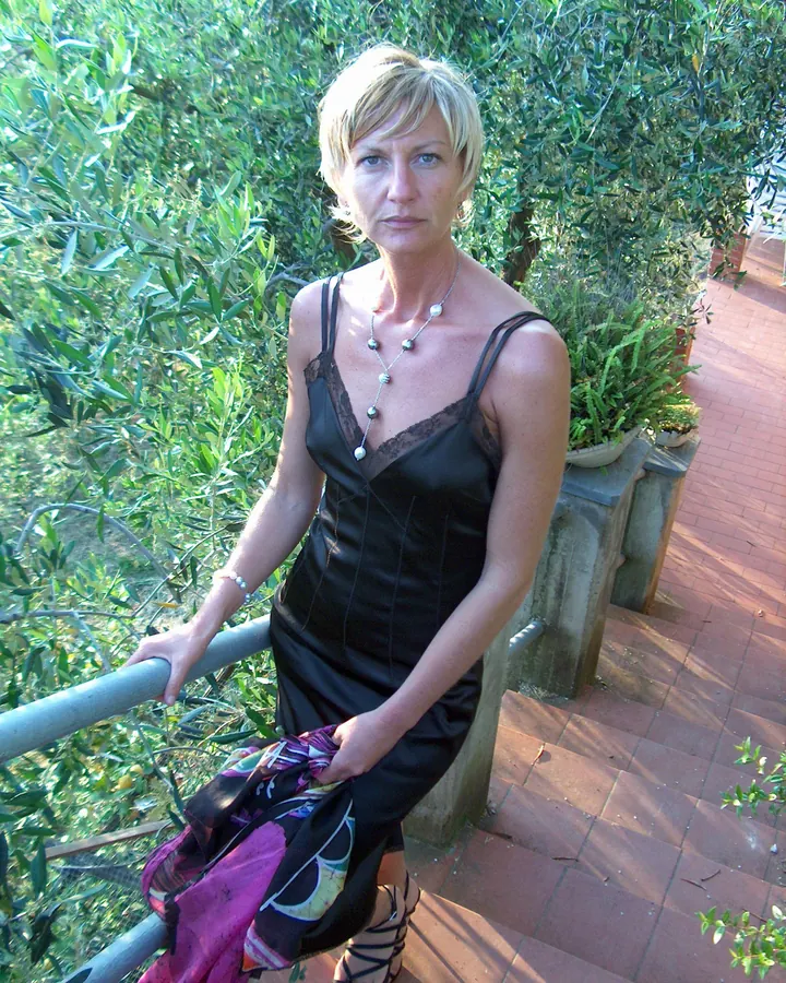 Femme mûre aux cheveux blonds, portant un haut noir, assise à une table de jardin, souriant légèrement.