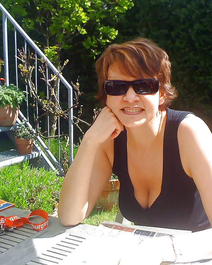 Femme mûre aux cheveux châtains courts, portant un haut blanc, appuyée contre un mur de pierres, souriant légèrement.