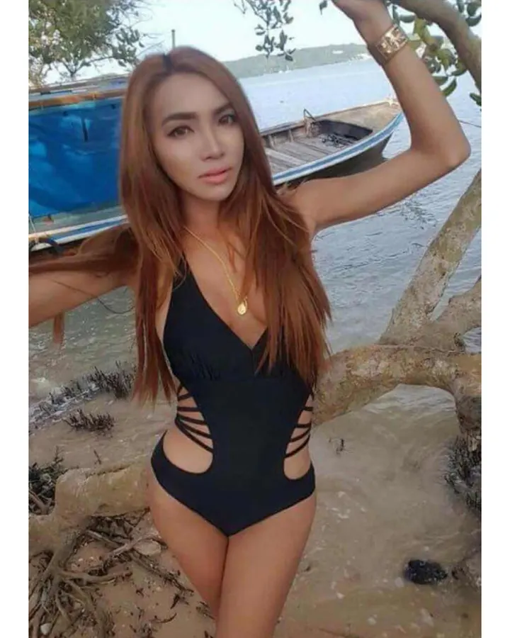Femme en maillot de bain noir, tenant une branche, près d’un bateau sur une plage.
