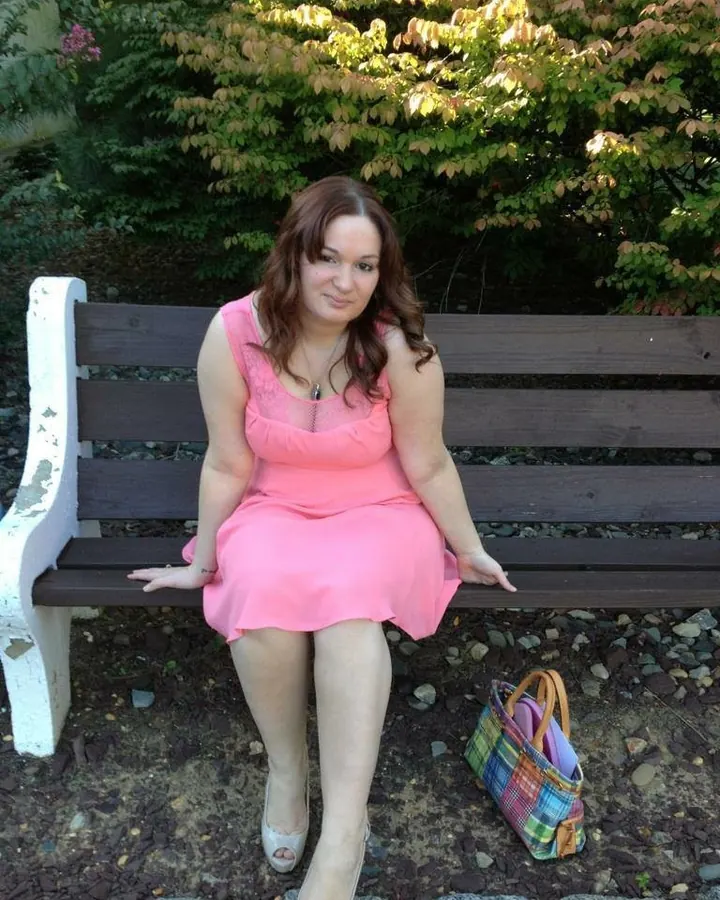 Femme en robe rose, assise sur un banc, sac coloré à côté, dans un jardin.